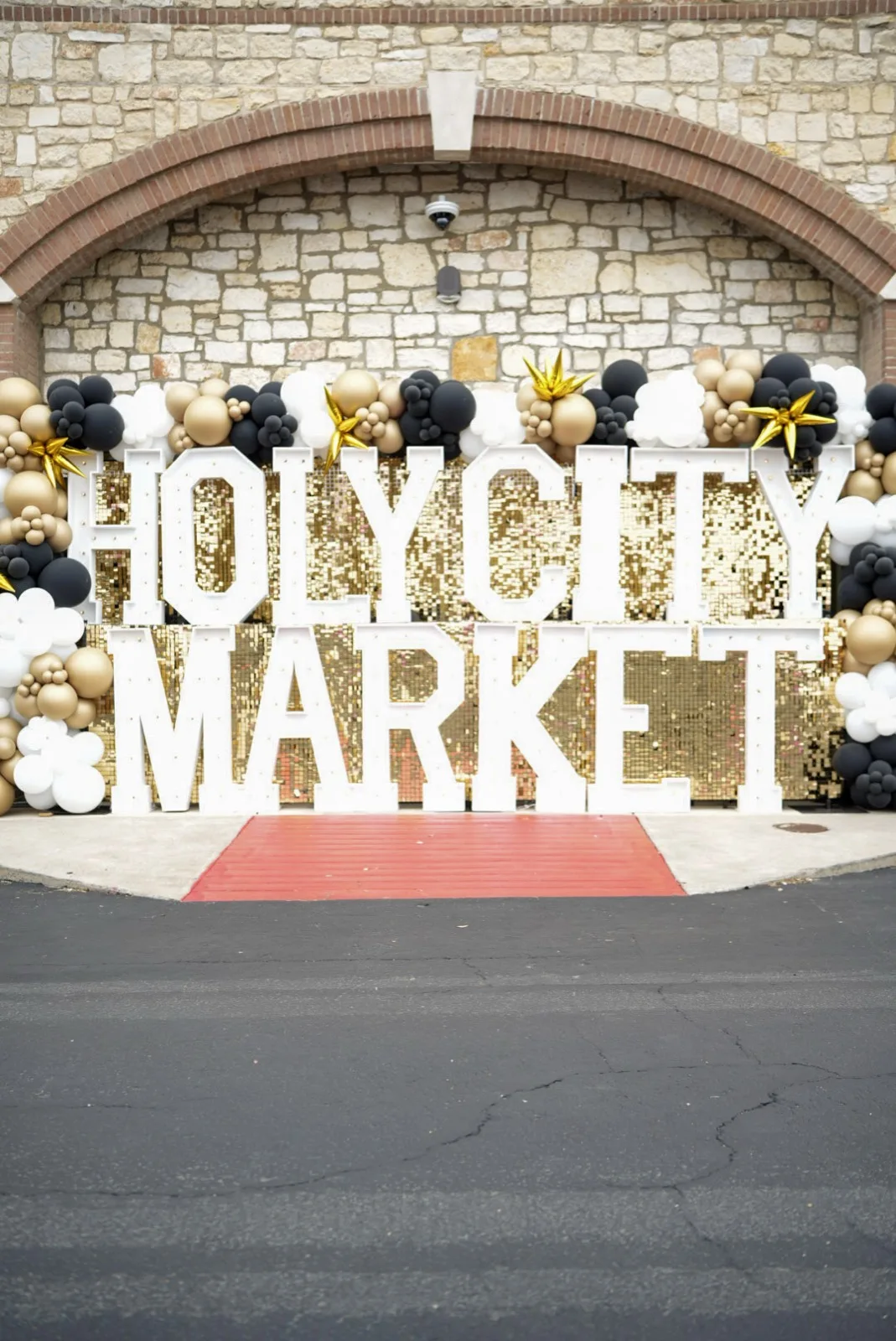 The illuminated HOLY CITY MARKET marquee letters against a gold sequin backdrop with black and gold balloon arch at the CBC West entrance.